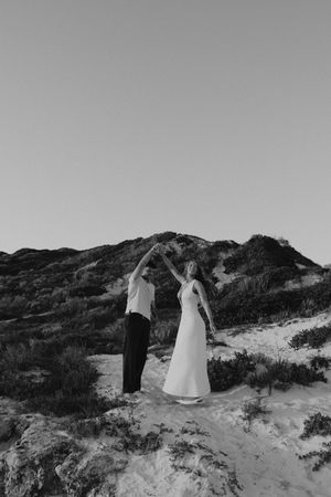 Black and white photo of a couple dancing on sandy dunes, woman in white dress, man in dark pants.