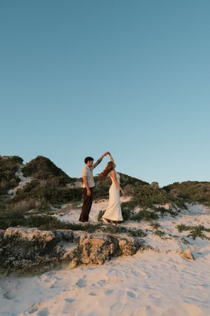Couple dancing on sandy beach dunes at sunset, woman in white dress being twirled by partner.