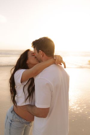 Romantic couple sharing a passionate kiss on a sunny beach at golden hour sunset.