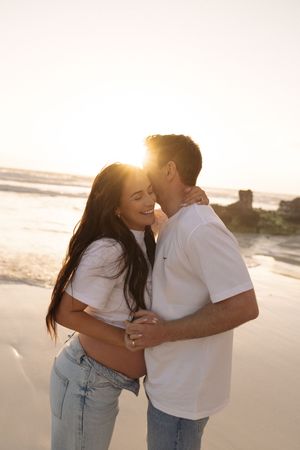 Couple embracing romantically on a beach at golden sunset, woman smiling as man kisses her cheek.