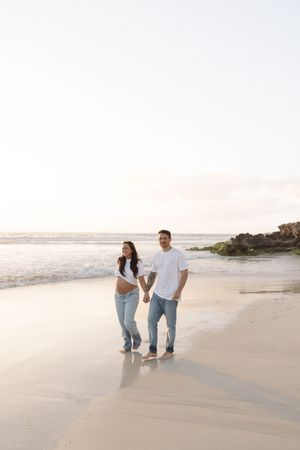 Pregnant couple holding hands and walking barefoot on a sandy beach at sunset in white tops and jeans.