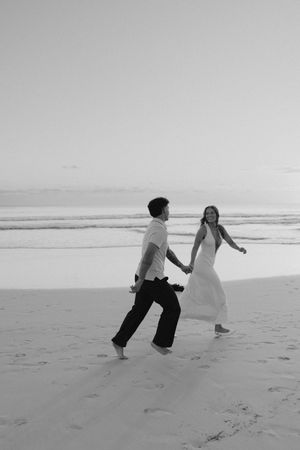 Black and white photo of joyful couple holding hands running along sandy beach at sunset.