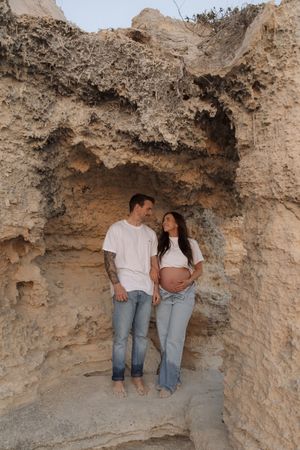 Couple in white tees and jeans posing in a sandstone cave alcove, woman showing baby bump in maternity photo.