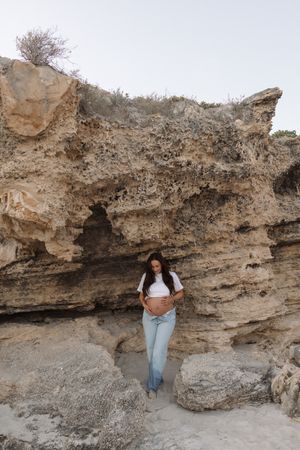 Pregnant woman in white crop top and jeans leaning against large sandy rock formation outdoors.