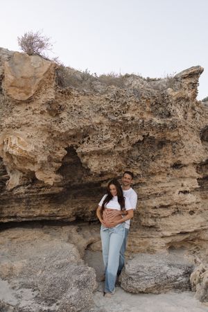 Pregnant couple embracing in front of large sandy limestone rock formation outdoors.