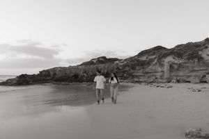 Black and white photo of a couple holding hands walking on a sandy beach with rocky cliffs at dusk.