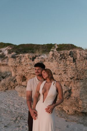 Couple embracing on sandy beach at sunset near rocky cliffs, woman in white halter dress, man in white polo shirt.