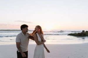 Laughing couple enjoying romantic beach sunset, woman in white dress with hair blowing in breeze.