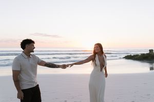 Couple holding hands facing each other on serene beach at pastel sunset by ocean shore.