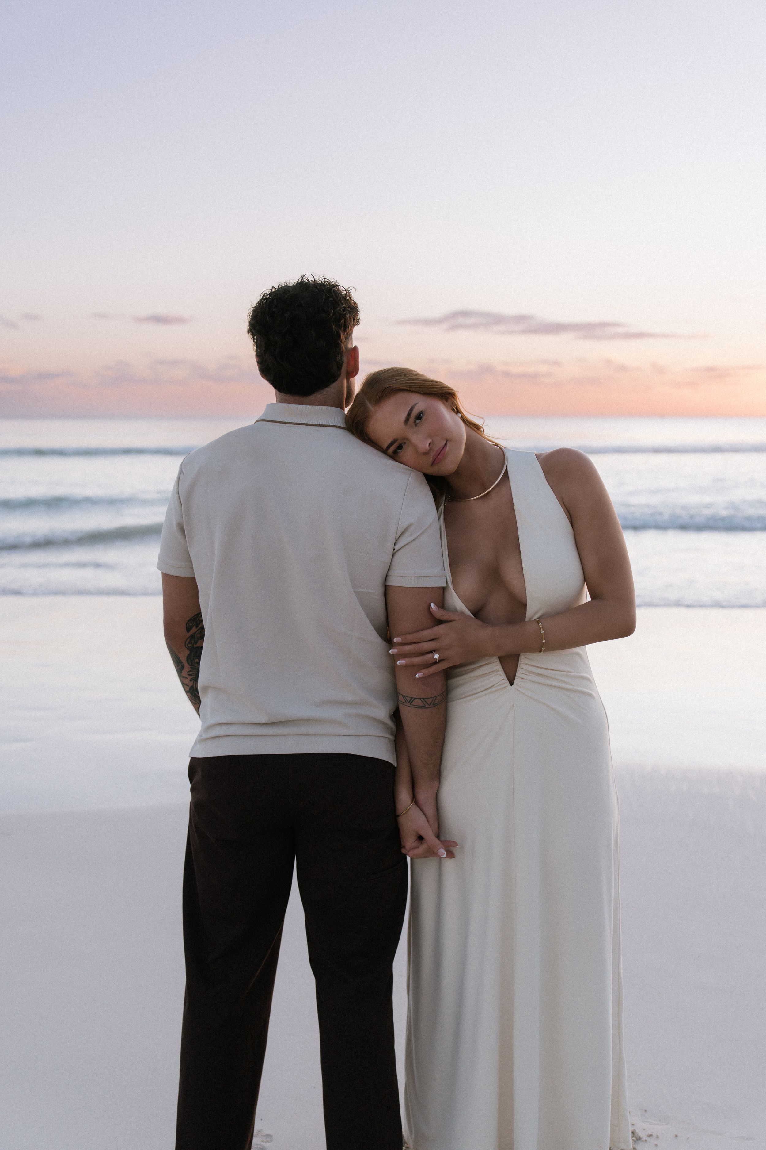 Woman in white dress leaning on mans shoulder at sunset beach, smiling softly at camera.