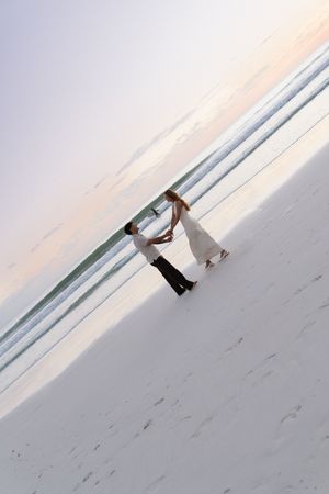 Aerial view of couple dancing together on white sand beach at pastel sunset by ocean.