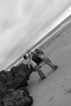 Black and white tilted angle shot of couple dancing playfully on sandy beach near rocks.