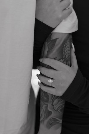 Black and white photo of a woman's hand with an engagement ring resting on a man's tattooed forearm.