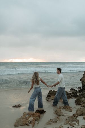 Couple holding hands walking along a sandy beach at sunset with rocky coastline.