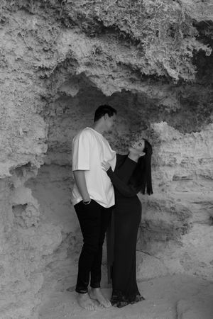 Black and white photo of a couple sharing a romantic moment in a sandstone cave alcove, woman in black dress gazing up at man.