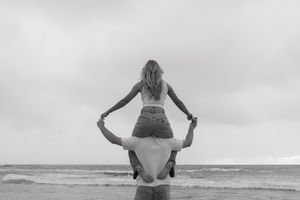 Black and white photo of woman sitting on mans shoulders at the beach, facing the ocean.