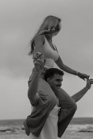 Black and white photo of a smiling man carrying a woman on his shoulders at the beach.