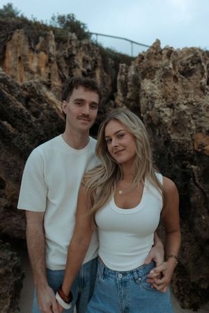 Couple in matching white tops and jeans posing in front of rocky coastal cliffs.
