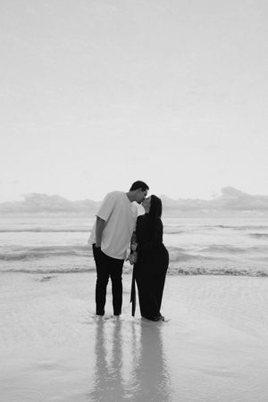 Black and white photo of a couple kissing on a beach shore with ocean waves in the background.