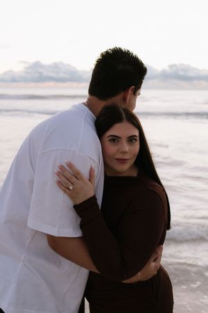 Couple embracing on beach at sunset, woman in brown outfit looks back at camera while man in white t-shirt faces ocean.