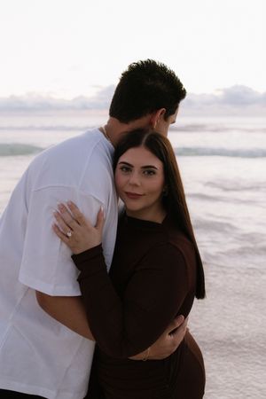 Couple embracing on a beach at sunset, woman in brown outfit looking at camera, man in white t-shirt.
