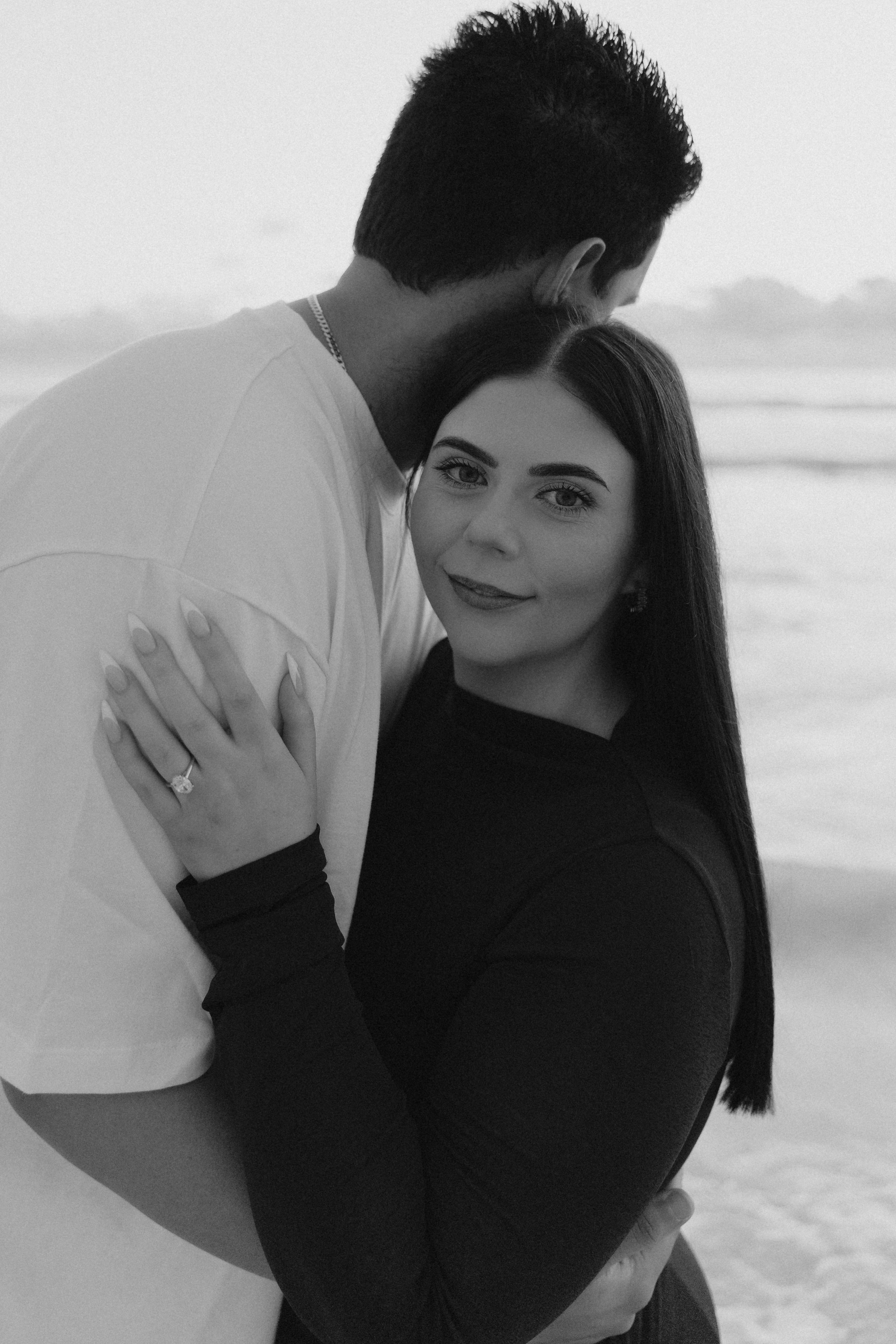 Black and white photo of a couple embracing on a beach, woman smiling at camera wearing an engagement ring.