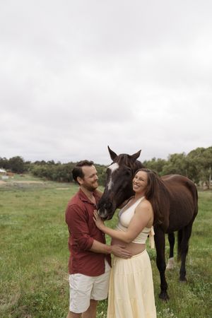 Couple smiling with dark horse in green field during maternity shoot.