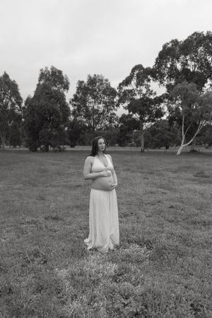 Black and white maternity portrait of pregnant woman in white dress outdoors.