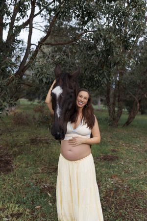 Pregnant woman in yellow skirt laughing beside dark horse outdoors.
