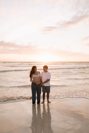 Pregnant couple standing on a sandy beach at sunset, holding baby bump together with ocean waves in the background.