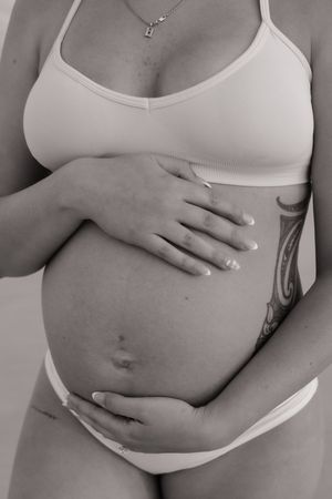 Black and white photo of a pregnant woman in white lingerie cradling her baby bump, with a tattoo visible on her side.