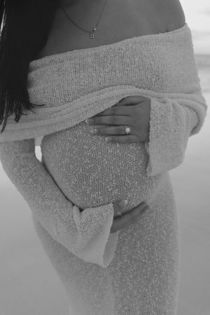 Black and white maternity photo of pregnant woman in off-shoulder knit dress cradling baby bump with diamond ring visible.
