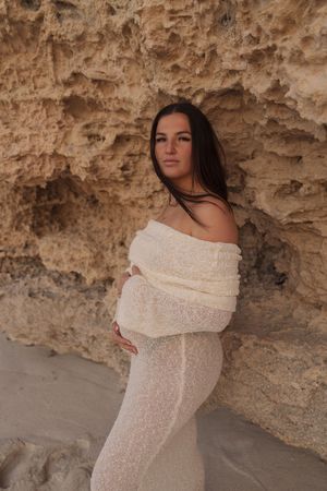 Pregnant woman in white lace off-shoulder gown poses against sandy rock wall, cradling baby bump in serene maternity photo.