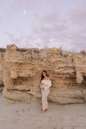 Pregnant woman in cream off-shoulder dress poses against sandy limestone cliffs on a beach at dusk.