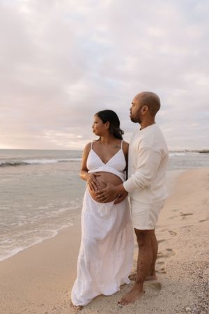 Pregnant couple in white outfits embracing on a beach at sunset, man tenderly holding woman's baby bump.