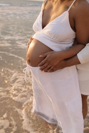 Pregnant woman in white dress at beach with partner's hands cradling baby bump near ocean waves.