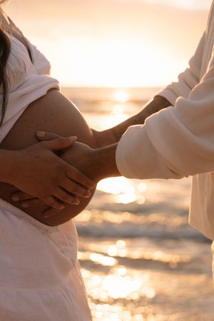 Pregnant woman and partner tenderly holding baby bump together on beach at golden sunset.