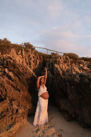 Pregnant woman in white dress poses between rocky cliffs at sunset beach, arm raised gracefully.
