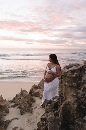 Pregnant woman in white dress cradling her belly on a rocky beach at sunset with pink clouds.