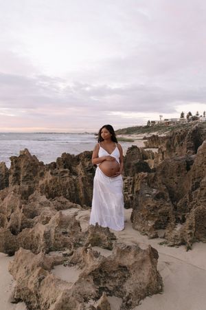 Pregnant woman in white dress cradling baby bump on rocky beach at sunset with ocean waves in background.