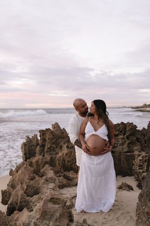 A couple shares a tender moment on a rocky beach at sunset, the woman in a white dress cradling her pregnant belly.