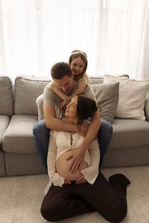 A happy family with a pregnant mother, father, and young daughter sharing a warm embrace in a cozy living room.