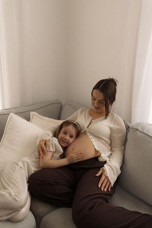 A young girl rests her head on her pregnant mother's belly while they relax together on a gray sofa.