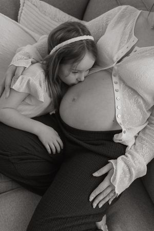 Black and white photo of a young girl kissing a pregnant woman's belly while sitting on a couch.