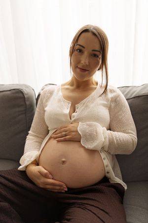 Pregnant woman in white top sitting on gray sofa, cradling her baby bump with both hands, smiling at camera.
