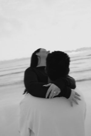 Black and white photo of a couple embracing on a beach, woman laughing joyfully while wearing an engagement ring.