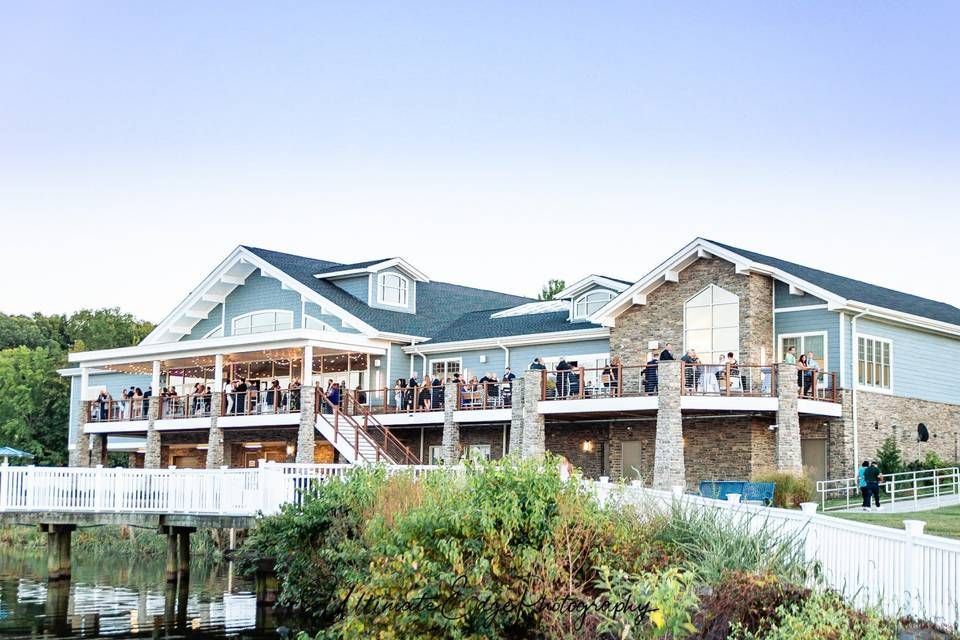 A scenic waterfront event venue with wrap-around decks and guests gathered for an outdoor celebration at sunset.