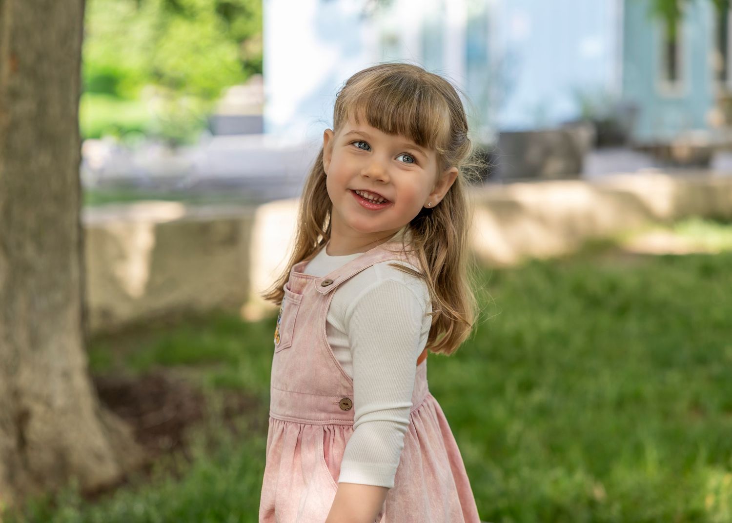 Young girl in pink dress and white top smiling outdoors in a green garden setting.