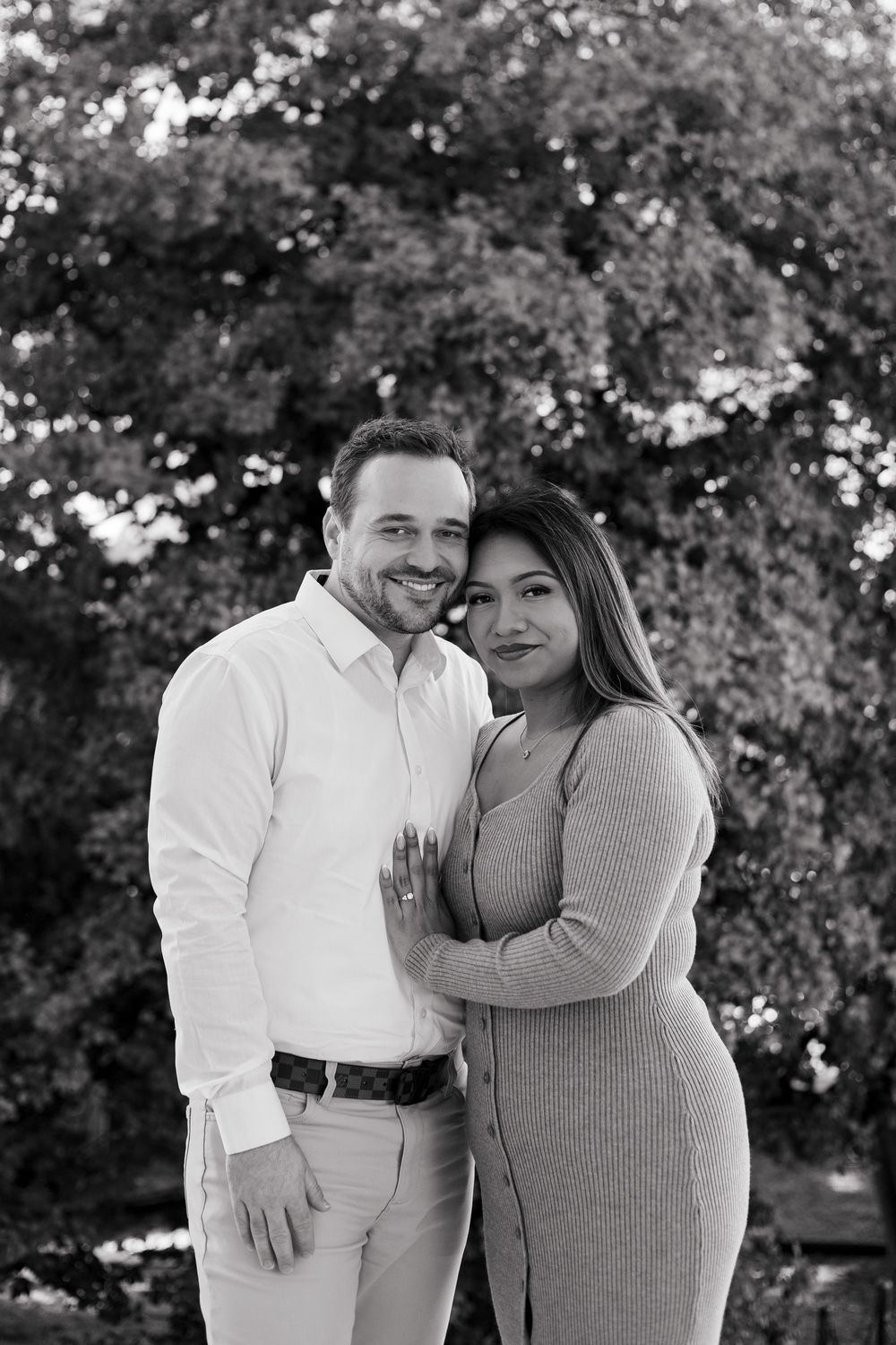 Black and white photo of a smiling couple embracing outdoors with lush trees in the background.