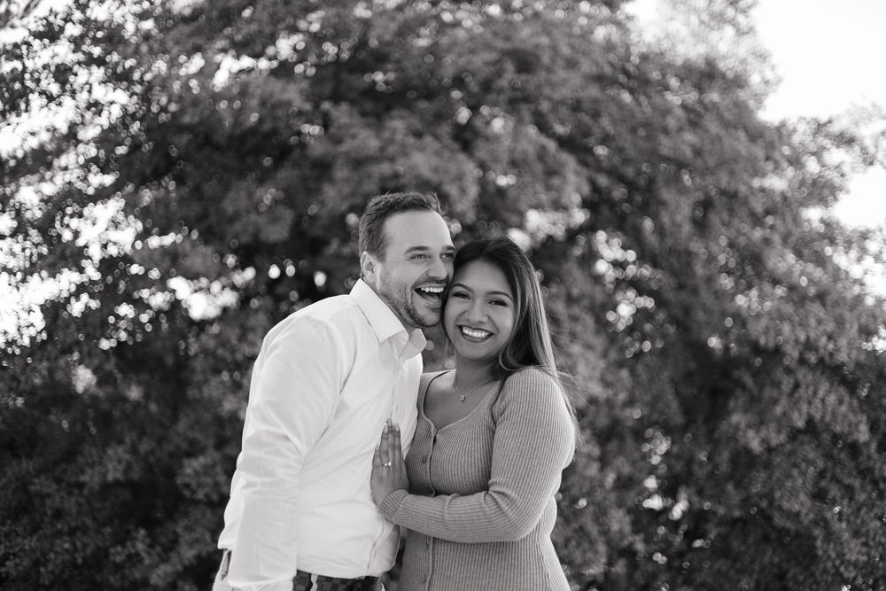 Black and white photo of a smiling couple embracing outdoors with lush trees in the background.